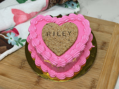 A small heart-shaped pink frosted cake or cookie on a gold-rimmed plate, placed on a wooden cutting board. The center is a brown heart-shaped cookie with the name 'RILEY' written on it.
